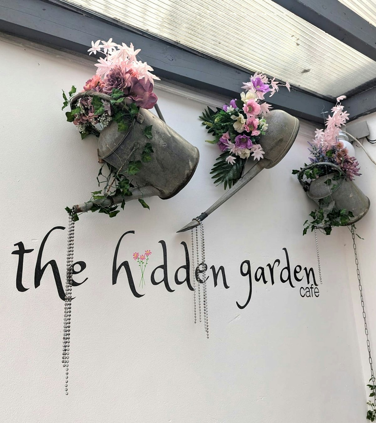 Three watering cans filled with flowers above a sign saying The Hidden Garden